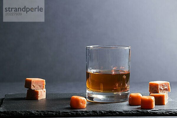 Glas bernsteinfarbener Whiskey mit Karamellbonbons auf einer schwarzen Schiefertafel aus Stein auf schwarzem Hintergrund. Seitenansicht  Kopierraum  selektiver Fokus