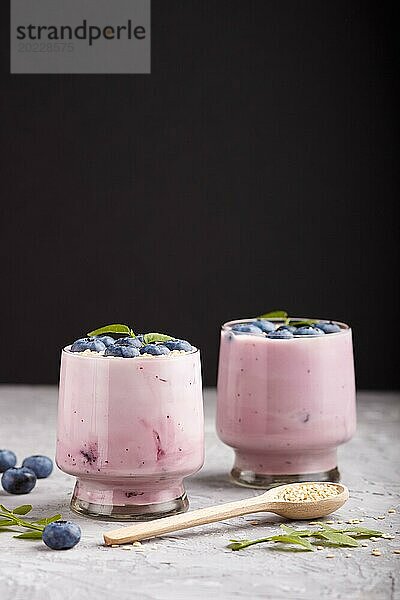 Joghurt mit Blaubeeren und Sesam im Glas und Holzlöffel auf grauem und schwarzem Hintergrund. Seitenansicht  Nahaufnahme