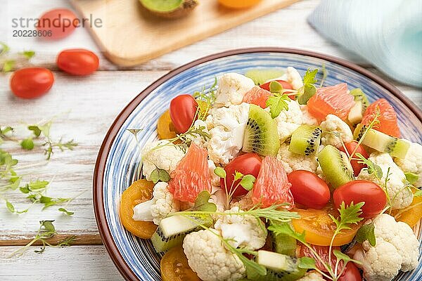 Vegetarischer Salat aus Blumenkohl  Kiwi  Tomaten  Mikrogrünsprossen auf weißem Holzhintergrund und blauem Leinentuch. Seitenansicht  Nahaufnahme  selektiver Fokus