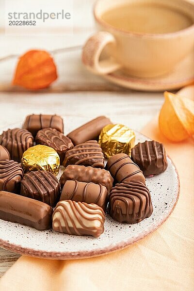 Schokoladenbonbons mit Kaffeetasse und Physalisblüten auf weißem Holzhintergrund und orangefarbenem Textil. Seitenansicht  Nahaufnahme  selektiver Fokus