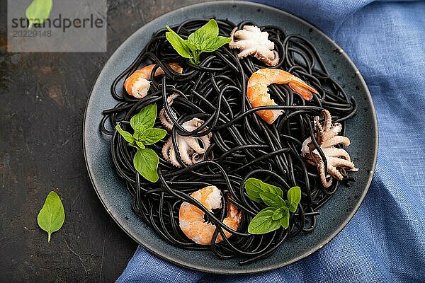 Schwarze Tintenfischnudeln mit Garnelen und kleinen Tintenfischen auf schwarzem Betonhintergrund und blauem Textil. Draufsicht  Nahaufnahme  selektiver Fokus