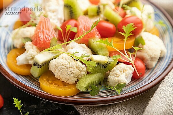 Vegetarischer Salat aus Blumenkohl  Kiwi  Tomaten  Mikrogrünsprossen auf schwarzem Betonhintergrund und Leinentuch. Seitenansicht  Nahaufnahme  selektiver Fokus