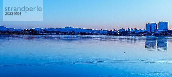 Die Skyline der Stadt spiegelt sich in der Morgendämmerung in Südkorea in ruhigem Wasser  das in blaue Farbtöne getaucht ist