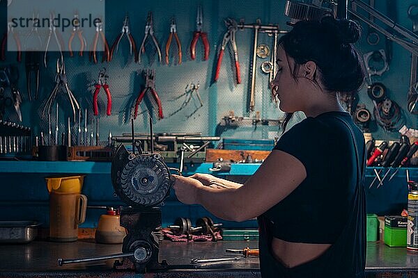 Gemischtrassige hispanische junge Mechanikerin  trägt schwarzen Overall  konzentriert sich auf ein Motorradteil in einer Werkstatt mit einem Werkzeugschrank  eine komplette Werkzeugtafel im Hintergrund mit Bokeh Effekt  traditionelle Männerberufe von gemischtrassiger Latino Frau