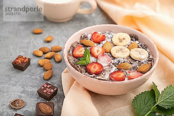 Schokoladen Cornflakes mit Milch  Erdbeeren und Mandeln in einer Keramikschale auf grauem Betonhintergrund und orangefarbenem Leinenstoff. Seitenansicht  Nahaufnahme  selektiver Fokus