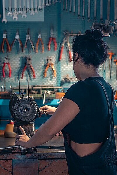 Weibliche hispanische Mechanikerin arbeitet an einer Werkbank in einer gut ausgestatteten Werkstatt von hinten  eine komplette Werkzeugtafel im Hintergrund mit Bokeh Effekt  traditionelle männliche Berufe von Mixed race latino woman