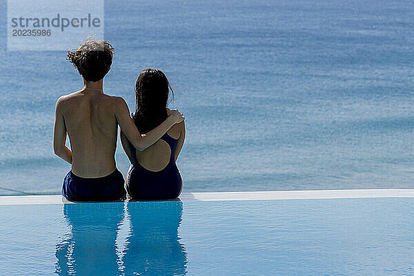 Junges Paar sitzt am Pool und blickt auf das Meer