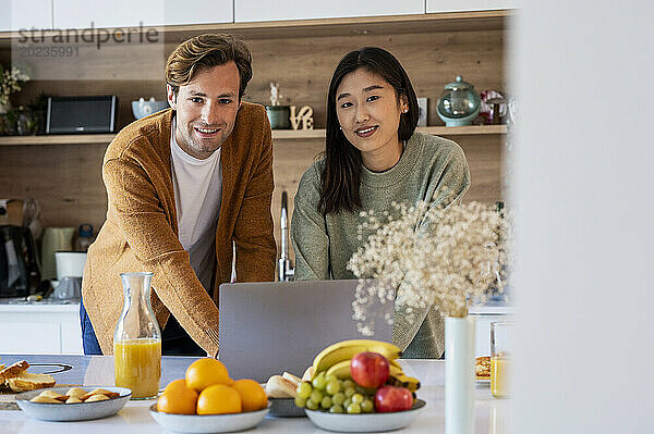 Erwachsenes Paar blickt in die Kamera  während es am Küchentisch einen Laptop benutzt