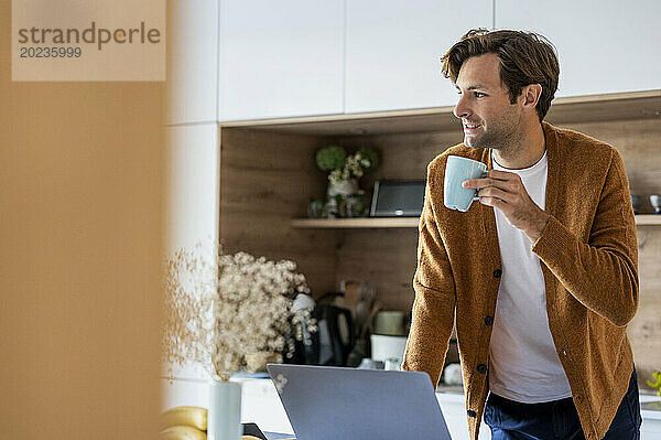 Nachdenklicher junger Mann trinkt eine Tasse Kaffee  während er in der Küche steht