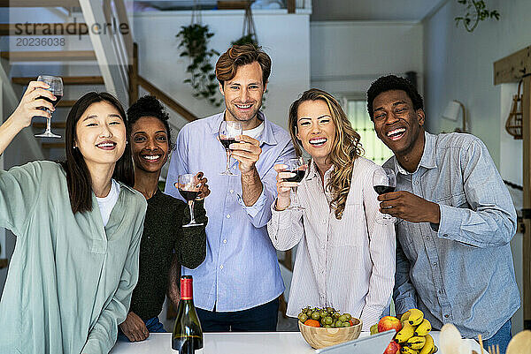 Eine Gruppe von Freunden blickt in die Kamera und prostet mit Weingläsern in der Küche zu