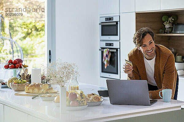 Erwachsener Mann kauft online am Laptop in der Küche ein