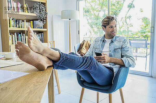 Junger erwachsener Mann ruht seine Füße aus  während er zu Hause mit dem Smartphone arbeitet