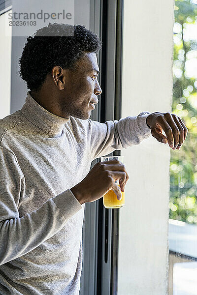 Seitenansicht eines erwachsenen Mannes  der sich am Fenster lehnt und Orangensaft trinkt