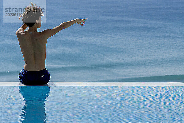 Junger Mann zeigt auf den Horizont  während er am Rand eines Swimmingpools am Meer sitzt