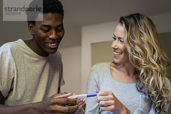 Happy couple smiling while holding pregnancy test in bedroom