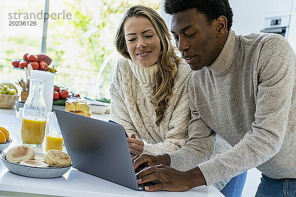 Adult man typing on laptop while leaning on kitchen counter with girlfriend