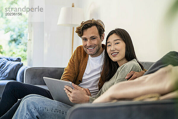 Erwachsenes Paar schaut in die Kamera  während es auf dem Sofa sitzt und ein digitales Tablet nutzt