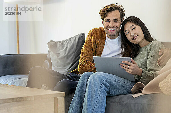 Erwachsenes Paar schaut in die Kamera  während es auf dem Sofa sitzt und ein digitales Tablet nutzt