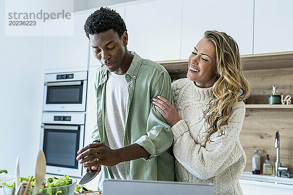 Erwachsene Frau umarmt ihren Freund beim Gemüseschneiden an der Küchentheke