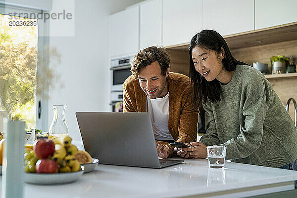 Erwachsenes Paar kauft online mit Laptop und Smartphone am Küchentisch ein