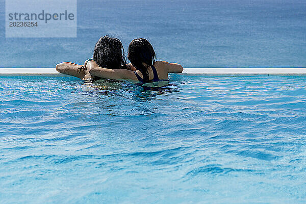 Rear view of two people embracing while looking at ocean from inside swimming pool
