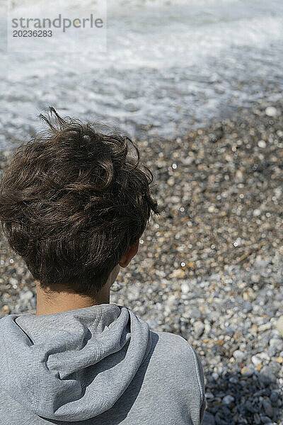 Rückansicht des Kopfes eines jungen Menschen mit Blick auf die Wellen am Meer in der Normandie