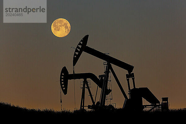 Silhouette zweier Pumpjacks mit leuchtend warmem Himmel und Vollmond  westlich von Airdrie; Alberta  Kanada