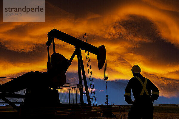 Silhouette eines männlichen Arbeiters und Pumpjacks vor einem farbenfrohen  dramatischen Sonnenuntergangshimmel mit warmen  leuchtenden Wolken  westlich von Airdrie; Alberta  Kanada