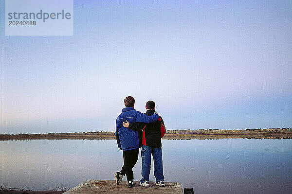 Freunde in einer Umarmung blicken in der Abenddämmerung auf einen ruhigen See  während sie auf einem Pfadfinderlager sind; Lincoln  Nebraska  Vereinigte Staaten von Amerika