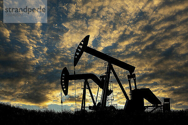 Silhouette eines Pumpjacks vor einem farbenfrohen  dramatischen Sonnenuntergangshimmel mit warmen  leuchtenden Wolken und blauem Himmel  westlich von Airdrie; Alberta  Kanada