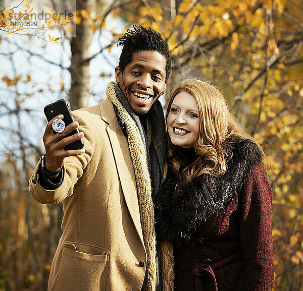 Nahaufnahme eines gemischtrassigen Paares  das gemeinsam ein Selfie macht  in die Handykamera lächelt und bei einem Familienausflug im Herbst in einem Stadtpark schöne Zeit miteinander verbringt; Edmonton  Alberta  Kanada