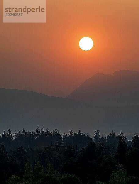 Orangefarbener Himmel in der Dämmerung über dunstigen Berggipfeln und silhouettiertem Wald; British Columbia  Kanada