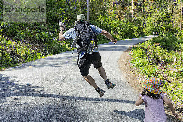 Rückansicht eines Vaters  der in die Luft springt  während seine Tochter dicht hinter ihm eine gepflasterte Straße im Lynn Valley Canyon entlangläuft; Nord-Vancouver  British Columbia  Kanada