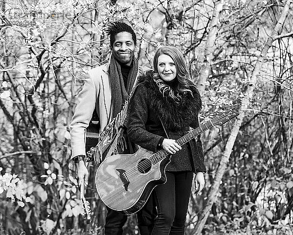 Porträt eines gemischtrassigen Ehepaares  das Akustikgitarren in der Hand hält und in die Kamera lächelt  während es bei einem Familienausflug im Herbst in einem Stadtpark schöne Zeit miteinander verbringt; Edmonton  Alberta  Kanada