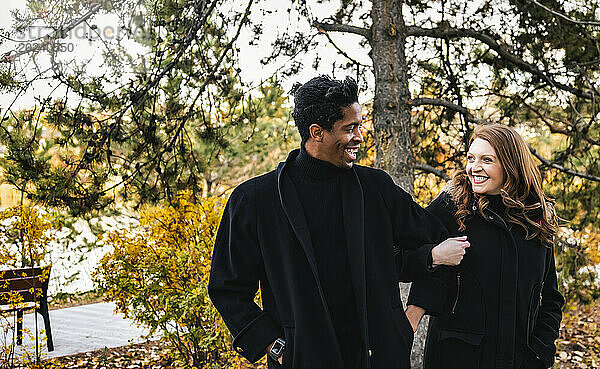 Ein Paar gemischter Abstammung  das sich gegenseitig anlächelt  während es durch den Wald spaziert und bei einem Familienausflug im Herbst in einem Stadtpark schöne Zeit miteinander verbringt; Edmonton  Alberta  Kanada
