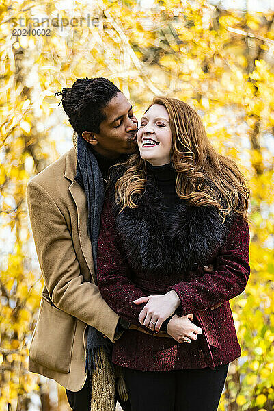 Ein gemischtrassiges Ehepaar umarmt seinen Mann und küsst seine Frau auf die Wange  während es bei einem Familienausflug im Herbst in einem Stadtpark schöne Zeit miteinander verbringt; Edmonton  Alberta  Kanada