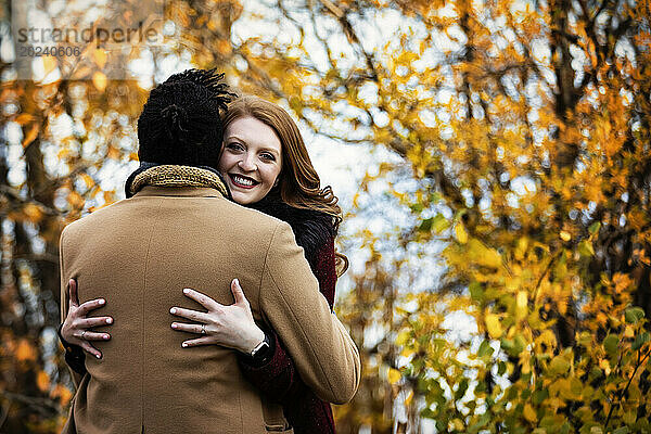 Blick von hinten auf ein Ehepaar gemischter Abstammung  das sich umarmt  während seine Frau in die Kamera lächelt  während es bei einem Familienausflug im Herbst in einem Stadtpark schöne Zeit miteinander verbringt; Edmonton  Alberta  Kanada