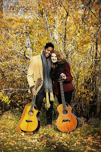Porträt eines gemischtrassigen Ehepaares  das Akustikgitarren in der Hand hält und in die Kamera lächelt  während es bei einem Familienausflug im Herbst in einem Stadtpark schöne Zeit miteinander verbringt; Edmonton  Alberta  Kanada
