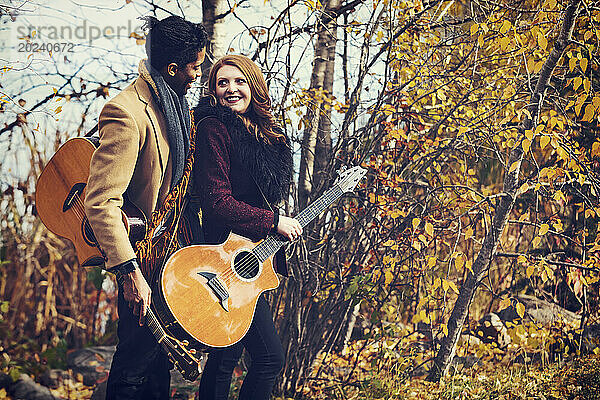 Porträt eines gemischtrassigen Ehepaares  das Akustikgitarren in der Hand hält und sich gegenseitig anlächelt  während es bei einem Familienausflug im Herbst in einem Stadtpark schöne Zeit miteinander verbringt; Edmonton  Alberta  Kanada