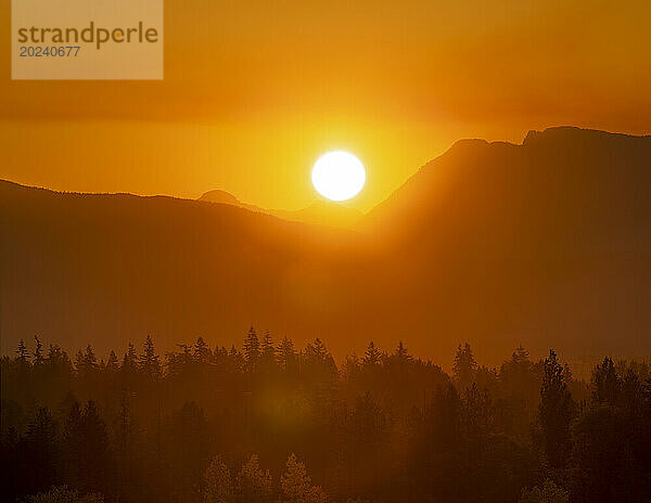 Eine helle Sonne mit orangefarbenem Himmel und goldenem Sonnenlicht  das in der Dämmerung über den Silhouetten der Berggipfel und des Waldes leuchtet; British Columbia  Kanada