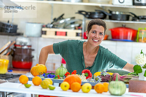 Lächelnde und entspannte Frau  mit einem Stapel Bio-Gemüse vor ihr und den Küchenutensilien im Hintergrund im Fokus.