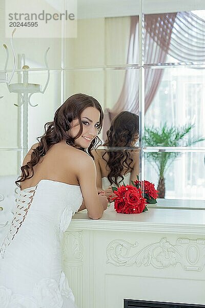 Attraktive lächelnde lockige Braut posiert vor einem Spiegel