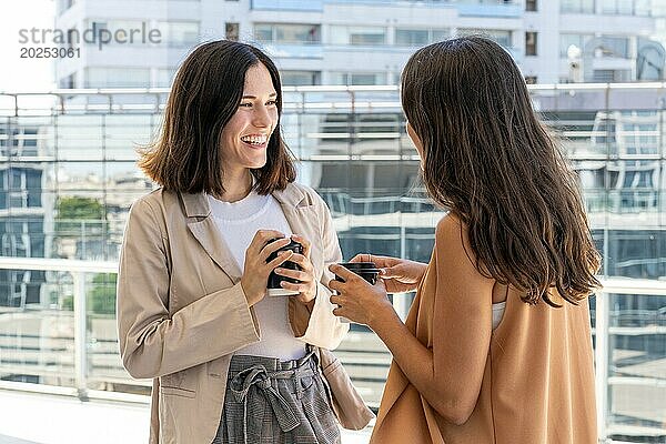 Zwei Geschäftsfrauen stehen vor einem Gebäude und genießen ihre Büropause. Sie lächeln und halten Tassen mit Kaffee in der Hand  was auf ein Gefühl der Kameradschaft und Freundschaft hindeutet