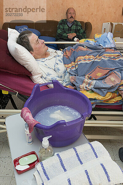 HAD (Hospitalisierung zu Hause). Betreuung einer älteren Patientin zu Hause durch eine Pflegekraft. Bassine posiert während der Toilette in der Nähe des Krankenbetts eines älteren und behinderten Patienten.
