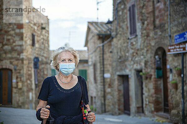 Ältere Frau trägt eine schützende Gesichtsmaske und hält einen Wanderstock  während sie in einer italienischen Stadt steht