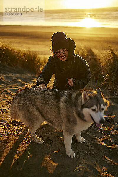 Lächelnde junge Frau streichelt Husky-Hund im Sand am Strand