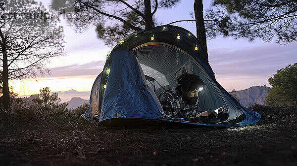 Mann mit Stirnlampe und Lesebuch im Zelt in der Nähe von Bäumen