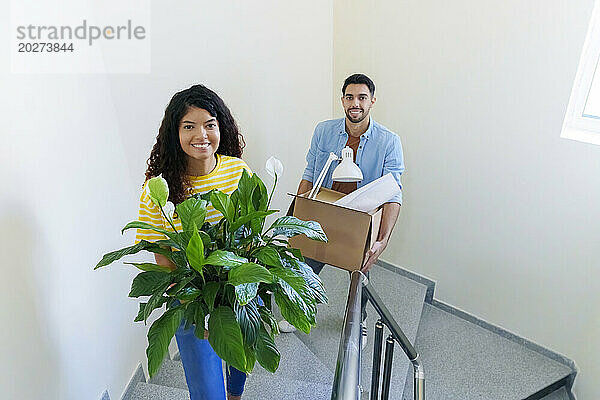 Lächelndes Paar trägt Kiste und Pflanze auf der Treppe zu Hause