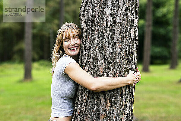 Lächelnde junge schöne Frau umarmt Baum im Wald