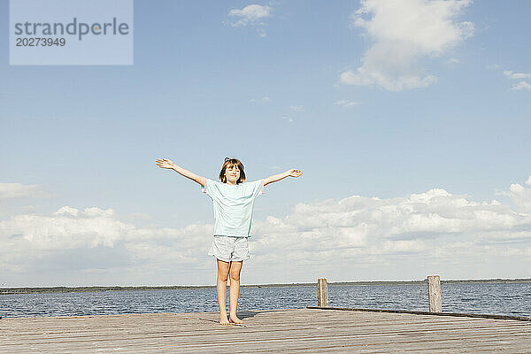 Mädchen steht mit ausgestreckten Armen auf einem Holzsteg am See